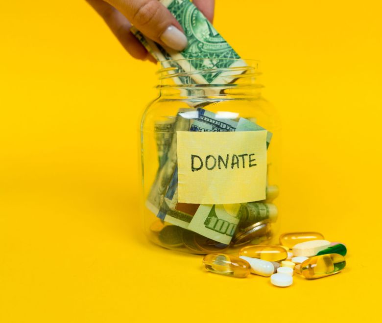 Donation jar with paper money and coins on yellow background. Pills and medicine. Womans hand putting dollar bill into the jar